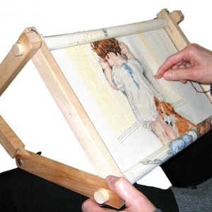 May include: Wooden embroidery hoop stand with a white fabric stretched across it. A person is working on an embroidery project with a needle and thread. The stand is adjustable and can be tilted to different angles.