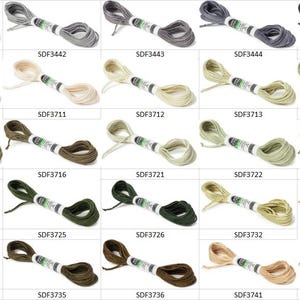 May include: A collection of twenty-five skeins of embroidery floss arranged in a grid, each featuring a unique color ranging from white, gray, and black to various shades of green, olive, and tan, with each skein labeled with a specific code.