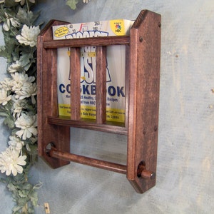 May include: A dark brown wooden wall-mounted cookbook holder with a towel rack. The holder has two shelves and a metal rod for hanging towels. The holder is mounted on a light blue wall.
