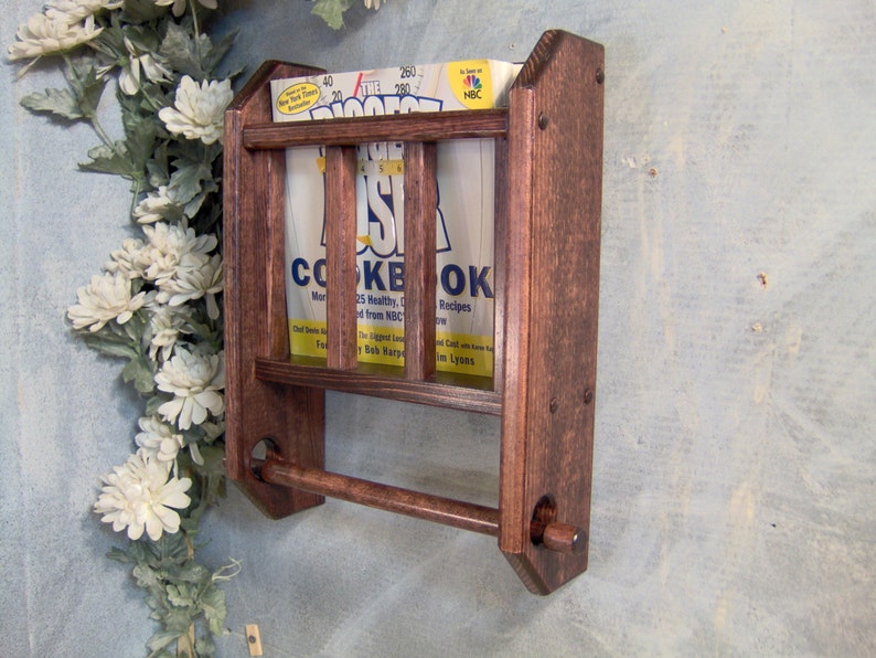 May include: A dark brown wooden wall-mounted cookbook holder with a towel rack. The holder has two shelves and a metal rod for hanging towels. The holder is mounted on a light blue wall.