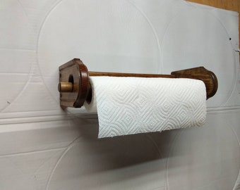 May include: A wooden paper towel holder with a brown finish. The holder is mounted to a wall and has a roll of white paper towels on it.