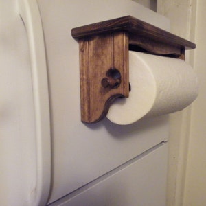 K&ouml;nnte beinhalten: Ein Papierrollenhalter aus Holz mit einer dunkelbraunen Beize. Der Halter ist an einer wei&szlig;en Wand montiert und hat eine Rolle wei&szlig;es Papier.