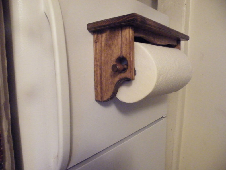 K&ouml;nnte beinhalten: Ein Papierrollenhalter aus Holz mit einer dunkelbraunen Beize. Der Halter ist an einer wei&szlig;en Wand montiert und hat eine Rolle wei&szlig;es Papier.