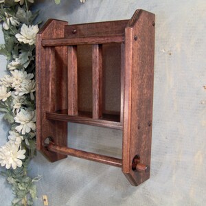 May include: A dark brown wooden wall shelf with a towel bar. The shelf has three compartments and is mounted on a wall.