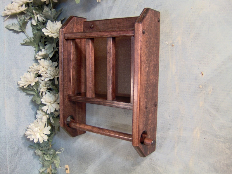 May include: A dark brown wooden wall shelf with a towel bar. The shelf has three compartments and is mounted on a wall.