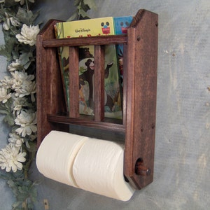 May include: A dark brown wooden wall-mounted shelf with two toilet paper rolls and a small bookcase with three children's books. The books have colorful covers with illustrations of animals and the text "Walt Disney" on one of the covers.