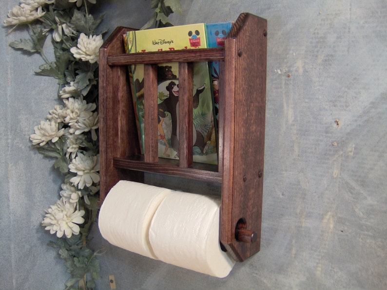 May include: A dark brown wooden wall-mounted shelf with two toilet paper rolls and a small bookcase with three children's books. The books have colorful covers with illustrations of animals and the text "Walt Disney" on one of the covers.