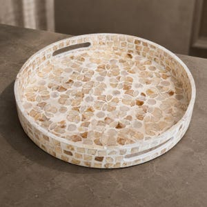 May include: A round serving tray featuring a mosaic pattern of light tan and cream-colored shell pieces arranged in floral motifs on the interior surface and a grid pattern on the exterior walls, complete with two integrated cutout handles.
