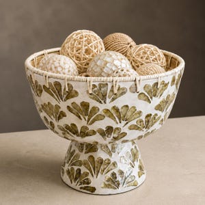 Puede incluir: Un cuenco con pedestal que presenta una base blanca con un patrón incrustado en forma de abanico de color verde oliva. El borde está envuelto en mimbre claro. El cuenco contiene varias esferas decorativas de ratán trenzado y concha clara.