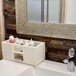 May include: White plastic bathroom organizer with five compartments, holding various bottles and tubes of toiletries. The organizer is sitting on a white countertop in front of a brown stone wall.