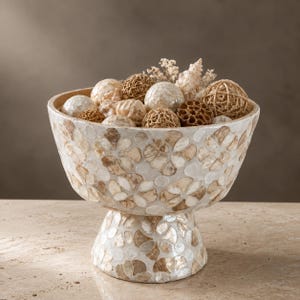 Könnte beinhalten: Eine Fußschale mit einem Mosaik-Außenbereich aus weißen und hellbraunen Muschelfragmenten in einem Blumenmuster. Die Schale ist mit einer Sammlung dekorativer Kugeln aus geflochtenem Rattan, glatten weißen Muscheln und getrockneten Pflanzenteilen gefüllt.
