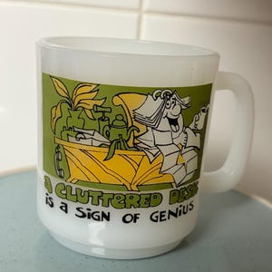 May include: White ceramic mug with a green and yellow graphic. The graphic depicts a cartoon desk scene with the text "A cluttered desk is a sign of genius."