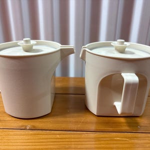 May include: Two matching cream-colored pitchers with lids and handles. The pitcher on the left has a spout, while the one on the right has a more rounded shape. Both are made of a matte material and sit on a wooden surface.