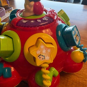 May include: A colorful, interactive baby toy with a red base and various attached shapes and buttons. Features a yellow star, green and blue accents, and a small cartoon figure on top. The toy is designed for early childhood development.