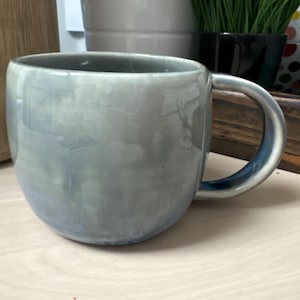 May include: A light blue ceramic mug with a rounded shape and a handle. The mug has a mottled, marbled glaze.