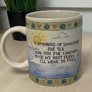 May include: A beige ceramic mug with a blue and green illustration of a sun, clouds, and a teacup. The mug has a quote that reads "A spoonful of sunshine for tea and you for company, ever my best friend I'll drink to thee."