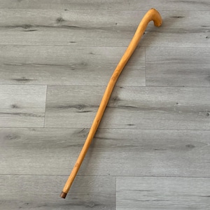 May include: A light brown wooden walking cane with a curved handle. The cane is angled across a gray wood-grain surface. The bottom of the cane has a small, dark brown metal tip.