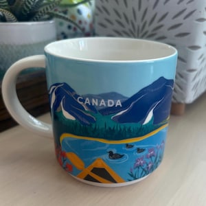 May include: A ceramic mug with a scenic illustration of Canada. The design features blue mountains, a lake with ducks, and the word "CANADA" in white. The mug has a white handle and interior.
