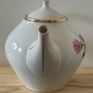 May include: White porcelain teapot with a gold rim and handle accents. The teapot features a floral design with pink roses. The lid has a small knob. The teapot is sitting on a wooden surface.