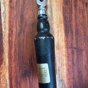 May include: A black bottle opener with a silver metal opener. The bottle opener has a label with the word "AWARDS" and the locations and years of awards. The bottle opener is on a wooden surface.