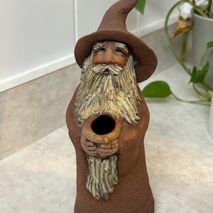 Peut inclure: Figurine de sorcier en céramique marron. Il porte un chapeau pointu et une longue barbe. Le sorcier tient un petit récipient ouvert. Les détails sont sculptés avec soin, lui donnant un aspect artisanal et rustique. Objet décoratif.