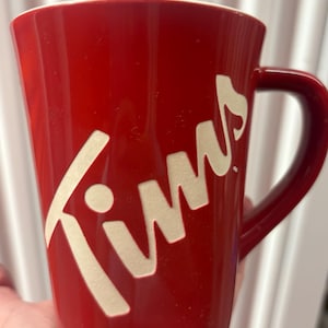 May include: A red ceramic mug with a white "Tims" logo. The mug has a curved handle and a slightly tapered shape. The interior of the mug is white.