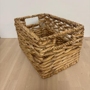 May include: A rectangular woven seagrass basket in a natural brown colour. The basket has a woven design with a handle on the top.