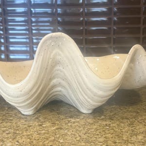 May include: A large, off-white ceramic clam shell-shaped bowl with a speckled surface. The bowl has a wavy, textured design and is suitable for decorative or serving purposes. The neutral colour and unique shape make it a versatile piece.