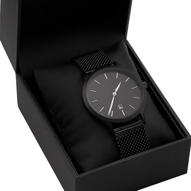 May include: A black wristwatch with a mesh band is displayed inside a black box. The watch has a black face with white hour markers and hands. The watch is a minimalist design, suitable for everyday wear.