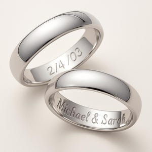 May include: Two polished silver wedding bands. One ring is engraved with "2/4/03", and the other with "Michael & Sarah". The rings have a smooth, rounded design, reflecting light. The bands are likely made of a precious metal.