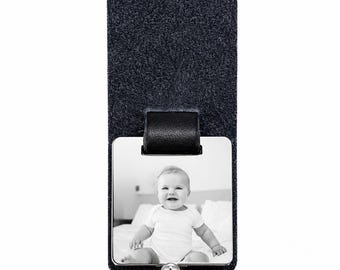 May include: A dark gray felt photo holder with a black leather strap and a silver-toned metal clasp. The holder displays a black and white photograph of a smiling baby. The top of the holder has a small hole for hanging.