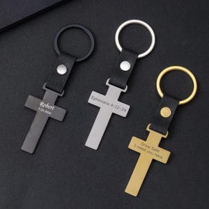 May include: Three cross-shaped keychains with black leather straps and metal rings. One is gunmetal gray with "Robert 5-20-2026", another is silver with "Ephesians 4:22-24", and the third is gold with "Drive Safe I need you here".