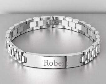 May include: A silver-colored bracelet with a watch-style band. The bracelet has a rectangular plate engraved with the name "Robert". The band is made of connected rectangular links.