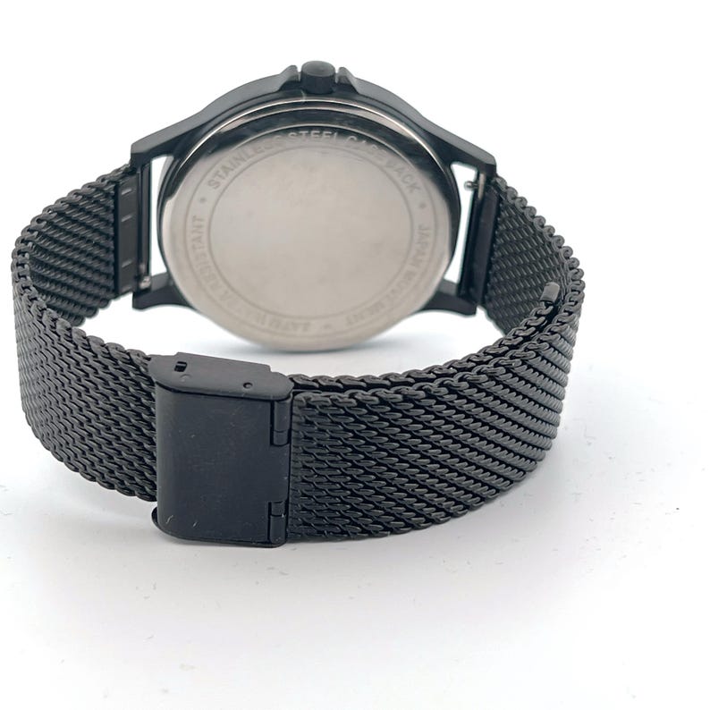 May include: A black wristwatch with a mesh band and a black watch face. The watch has a stainless steel back and a Japanese movement. The band is secured with a black clasp.