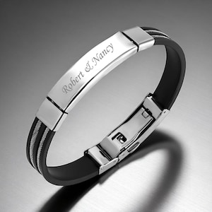 May include: A black and silver bracelet with the engraved text "Robert & Nancy." The bracelet features a black rubber band, a silver metal plate with the engraved text, and a silver clasp. The bracelet has a modern, sleek design.