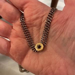 Puede incluir: Un collar de cadena de oro con un colgante redondo que presenta una piedra preciosa roja en el centro.