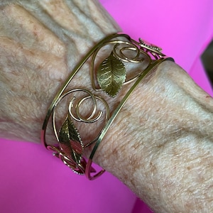 Puede incluir: Brazalete de puño dorado con diseño de hojas y espirales. El brazalete calado tiene dos bandas conectadas por elementos decorativos, incluyendo formas de hojas detalladas. El brazalete se lleva en la muñeca.