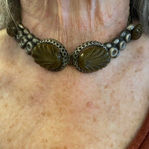 Op de afbeelding: Een choker-halsketting met twee grote, ovale, olijfbruine gesneden hars-cabochons in sierlijke zilverkleurige metalen monturen. De ketting bestaat uit kleine, ronde, getextureerde zilverkleurige metalen kralen en extra olijfbruine harsaccenten.