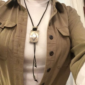 May include: A silver bolo tie with a rectangular pendant featuring a diamond-shaped design. The tie has a black cord and is worn over a white turtleneck and a tan jacket.