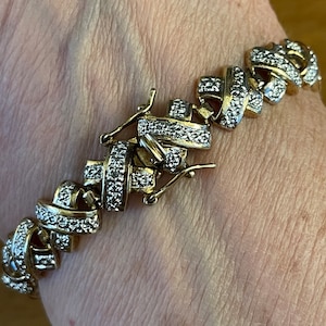 May include: A gold-toned bracelet with a repeating knot design, encrusted with small, sparkling stones. The bracelet is secured with a clasp. The jewelry is displayed on a skin-toned background.