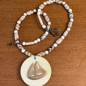 May include: A cream-coloured pendant necklace with a sailboat design. The beaded chain features alternating white and brown beads. The clasp is a silver-toned toggle. A unique nautical-themed accessory, perfect for adding a touch of the sea.