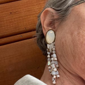 May include: An oval-shaped earring with a large, iridescent stone set in a bronze-colored frame. White and iridescent beads dangle below, creating a layered, fringe-like effect. The earring is a statement piece.