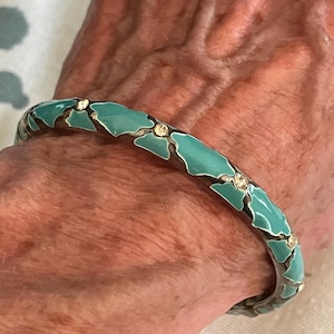 May include: A silver-toned bangle bracelet with a turquoise enamel floral design and small clear crystals. The bracelet is worn on a wrist and has a vintage or antique appearance.