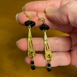 May include: A pair of dangle earrings featuring gold-tone metal triangular components with cut-out details, accented by black beads at the top and bottom. The earrings are suspended from gold-tone metal ear wires and have a textured finish.