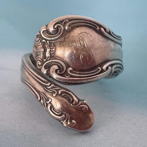 May include: A silver spoon ring with an ornate design. The ring features a decorative, engraved band with a large letter "B" in the center. The ring is made from a spoon that has been bent into a ring shape.