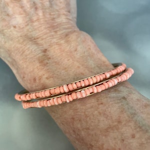May include: Two coral-colored beaded bracelets. The beads are strung on a thin, gold-colored metal band. The bracelets are stacked on top of each other. The bracelets are on a light skin tone.