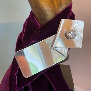 May include: A silver-toned brooch with a geometric design, featuring a large, clear gemstone. The brooch is set against a deep purple velvet fabric, creating a striking contrast. The design is modern and eye-catching.