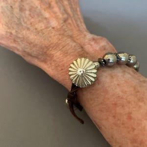 Pode incluir: Pulseira prateada com um fecho grande e decorativo em forma de sol com um centro quadrado. A pulseira inclui esferas prateadas e é presa a uma faixa de couro marrom escuro. A pulseira foi projetada para ser usada ao redor do pulso.