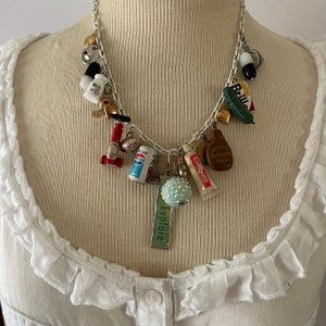 May include: A silver-toned chain necklace adorned with a variety of miniature charms. Charms include a pill, a miniature Pepsi can, a tube of Colgate toothpaste, a pickle, and a charm that says "explore". The necklace is displayed on a white background.