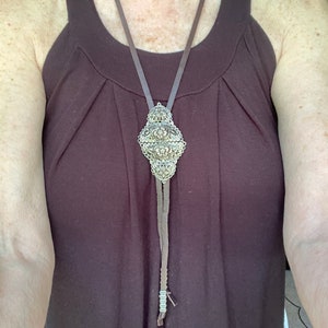 Può includere: Una cravatta a bolo decorativa con un pendente in filigrana argentata dettagliata. Il pendente è sospeso a un cordino in pelle marrone. La cravatta a bolo è indossata sopra una canotta marrone scuro.